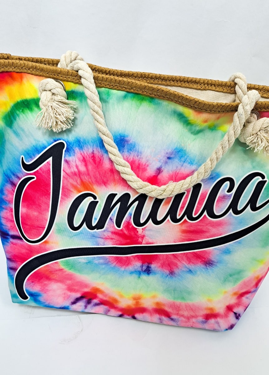 A colorful tie-dye bag with the word Jamaica written across it in bold, black script. The bag features rope handles and vibrant swirls of pink, yellow, blue, and green.