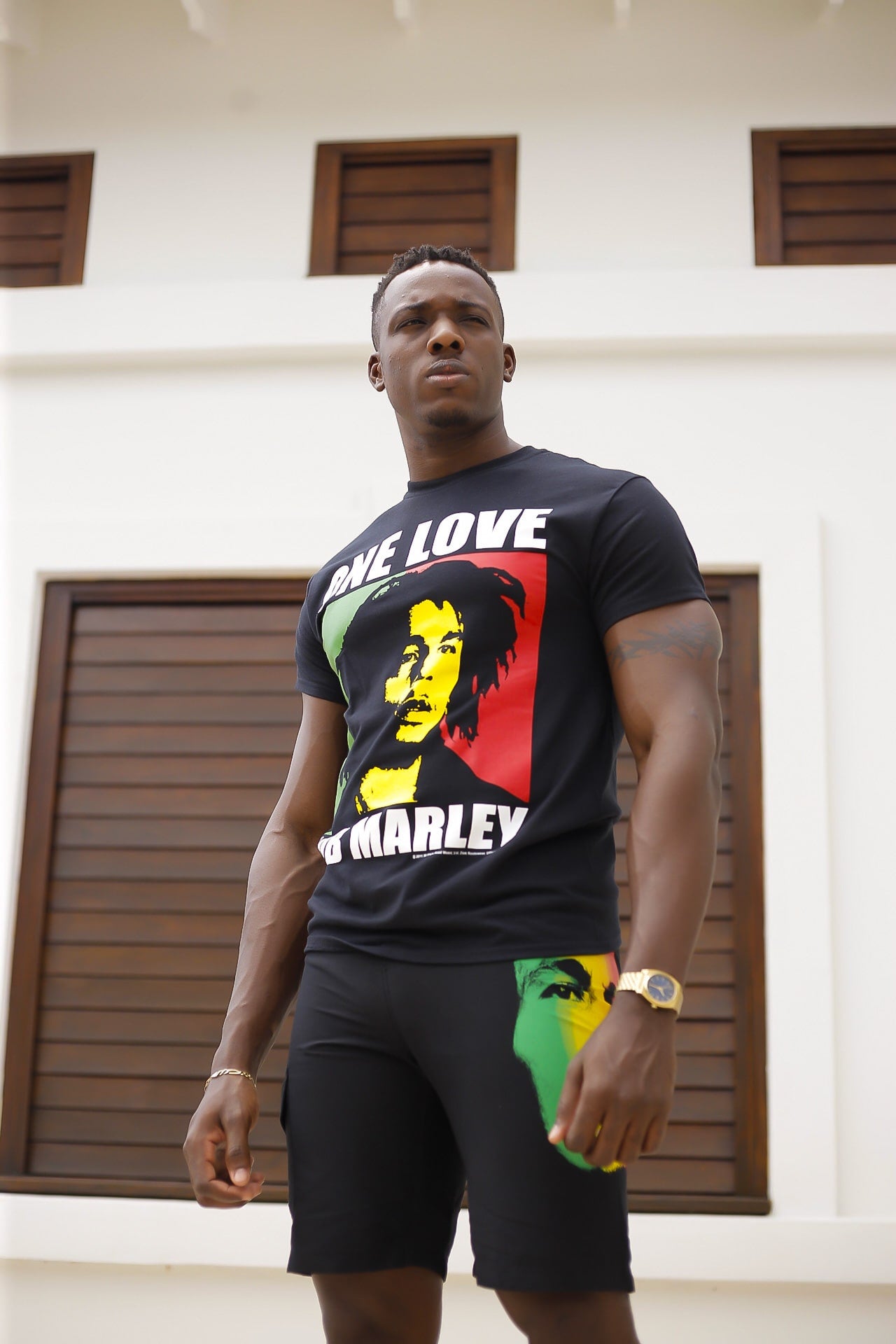 A man stands confidently wearing a shirt and shorts with a vibrant Bob Marley design, against a modern building with wooden shutters.